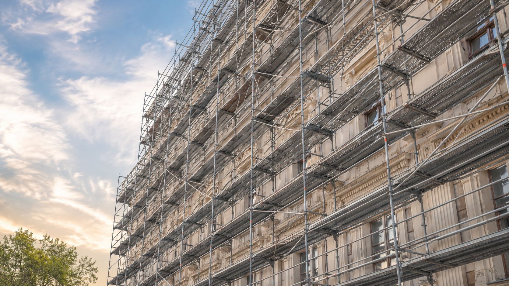 En historisk bygning under omfattende facaderenovering dækket af et stort stillads mod en flot aftenhimmel.