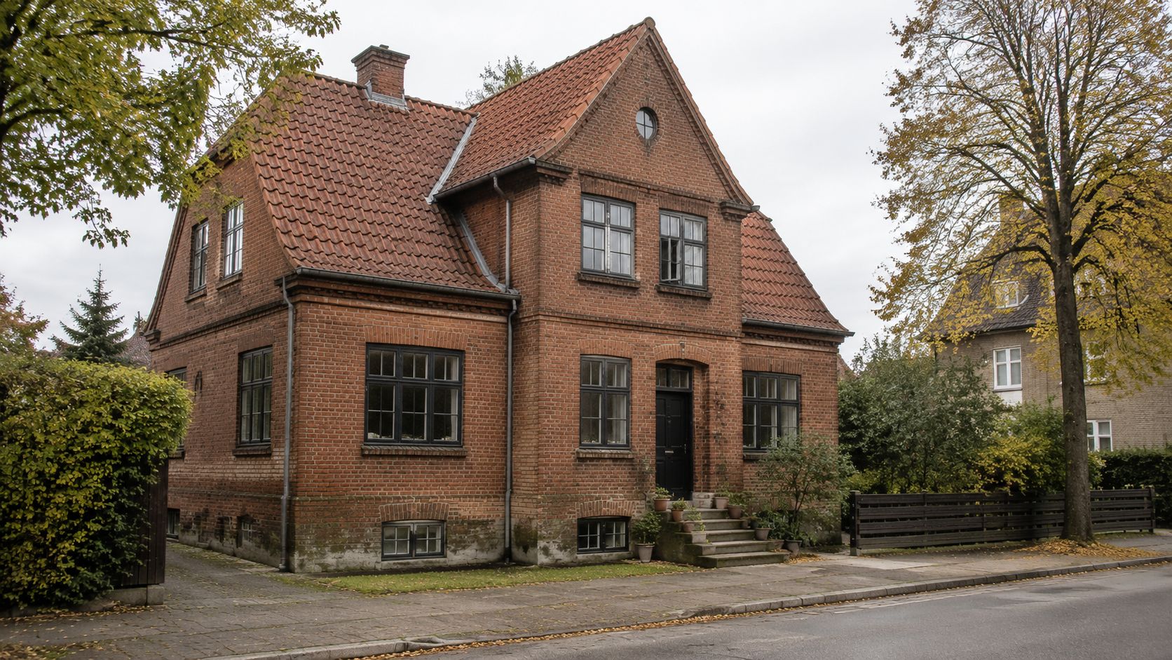 En ældre, klassisk murstensvilla med tegltag beliggende ved en vej med træer i efterårsfarver i baggrunden.